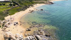 Ein Küstenstrand mit klarem Wasser, felsigem Ufer und einigen Gebäuden im Hintergrund.