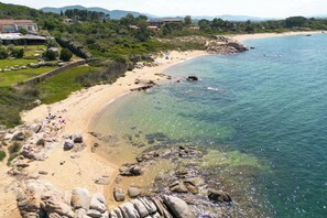 Ein Küstenstrand mit klarem Wasser, felsigem Ufer und einigen Gebäuden im Hintergrund.