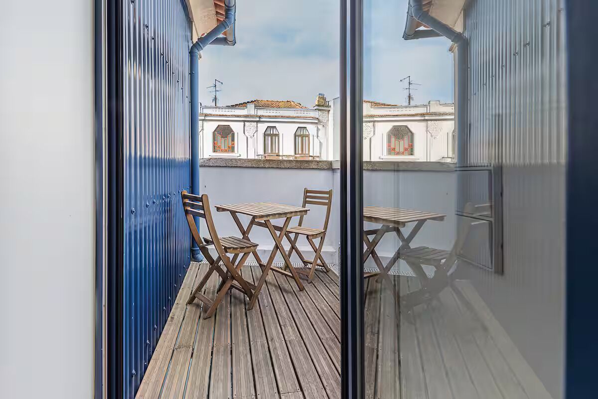 Ein Balkon mit Holzboden und einem Tisch mit Stühlen, mit Blick auf ein Gebäude mit weißen Wänden und Fenstern.