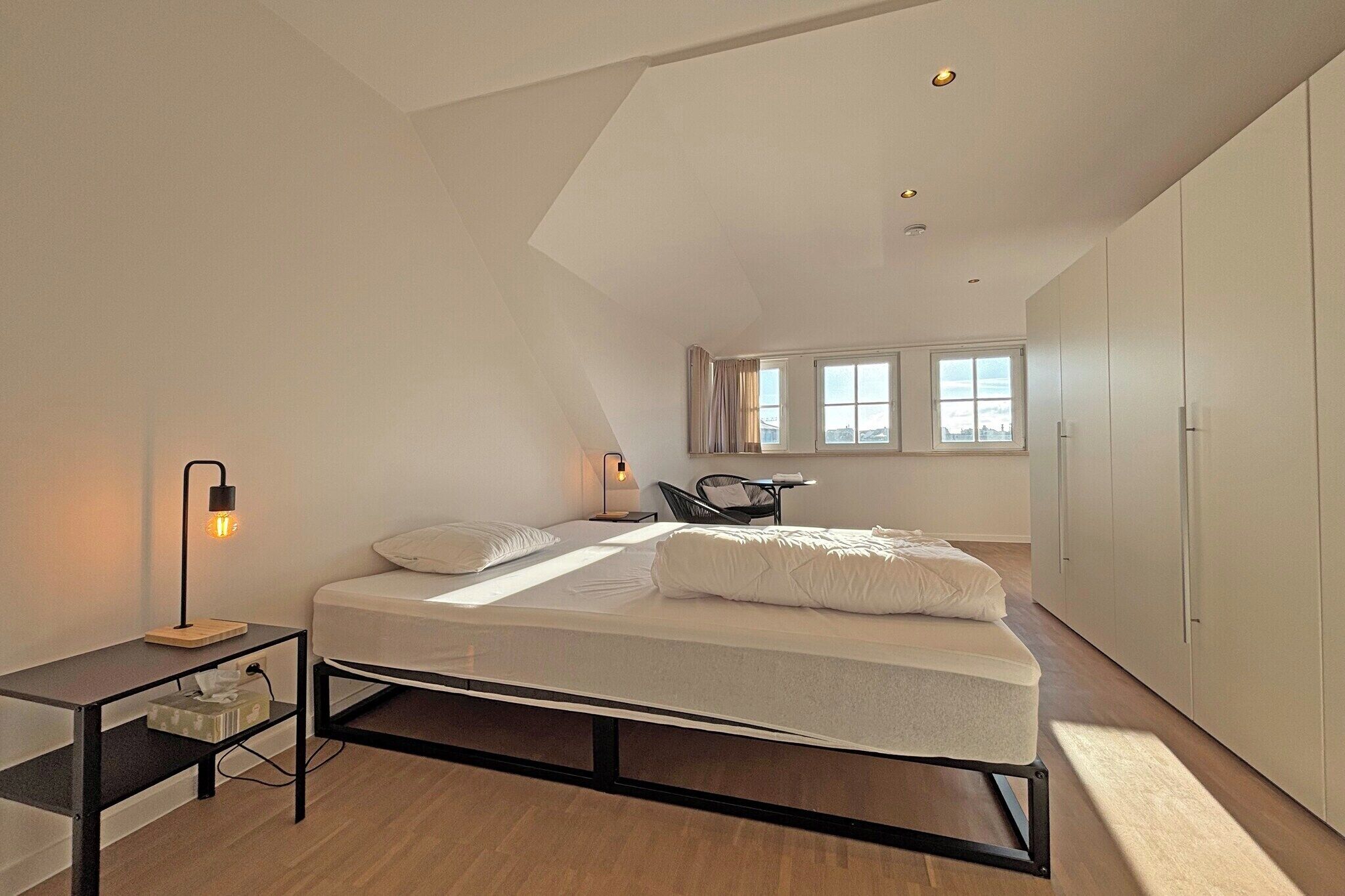 Een moderne slaapkamer met een bed, nachtkastje, lamp en uitzicht op de stad door ramen.