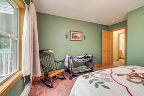 Um quarto com uma cadeira de balanço, um cercadinho para bebê, uma porta e uma janela grande.