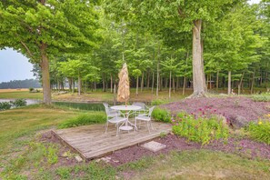 Um deck de madeira com mesa e cadeiras, rodeado por um jardim com diversas plantas e um lago.