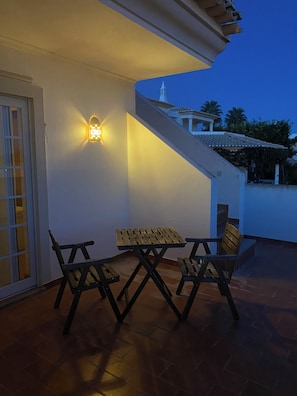An outdoor patio with a wooden table and chairs, a wall-mounted light fixture, and a view of a building with a tiled roof in the background. - Villa Med - private pool, garden, barbecue, air conditioning, and ocean view (Olhos de Água)
