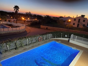 A swimming pool with a surrounding fence and a lifebuoy, set against a backdrop of buildings and a sunset sky. - Villa Med - private pool, garden, barbecue, air conditioning, and ocean view (Olhos de Água)