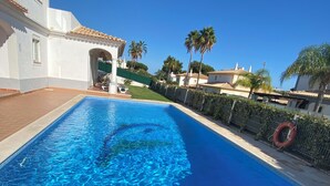 Ein Swimmingpool mit klarem, blauem Wasser, umgeben von einem weißen Gebäude mit einem Ziegeldach, Palmen und einer Hecke.