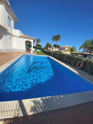 A swimming pool with clear blue water, surrounded by a white building with a tiled roof, palm trees, and a hedge. - Villa Med - private pool, garden, barbecue, air conditioning, and ocean view (Olhos de Água)