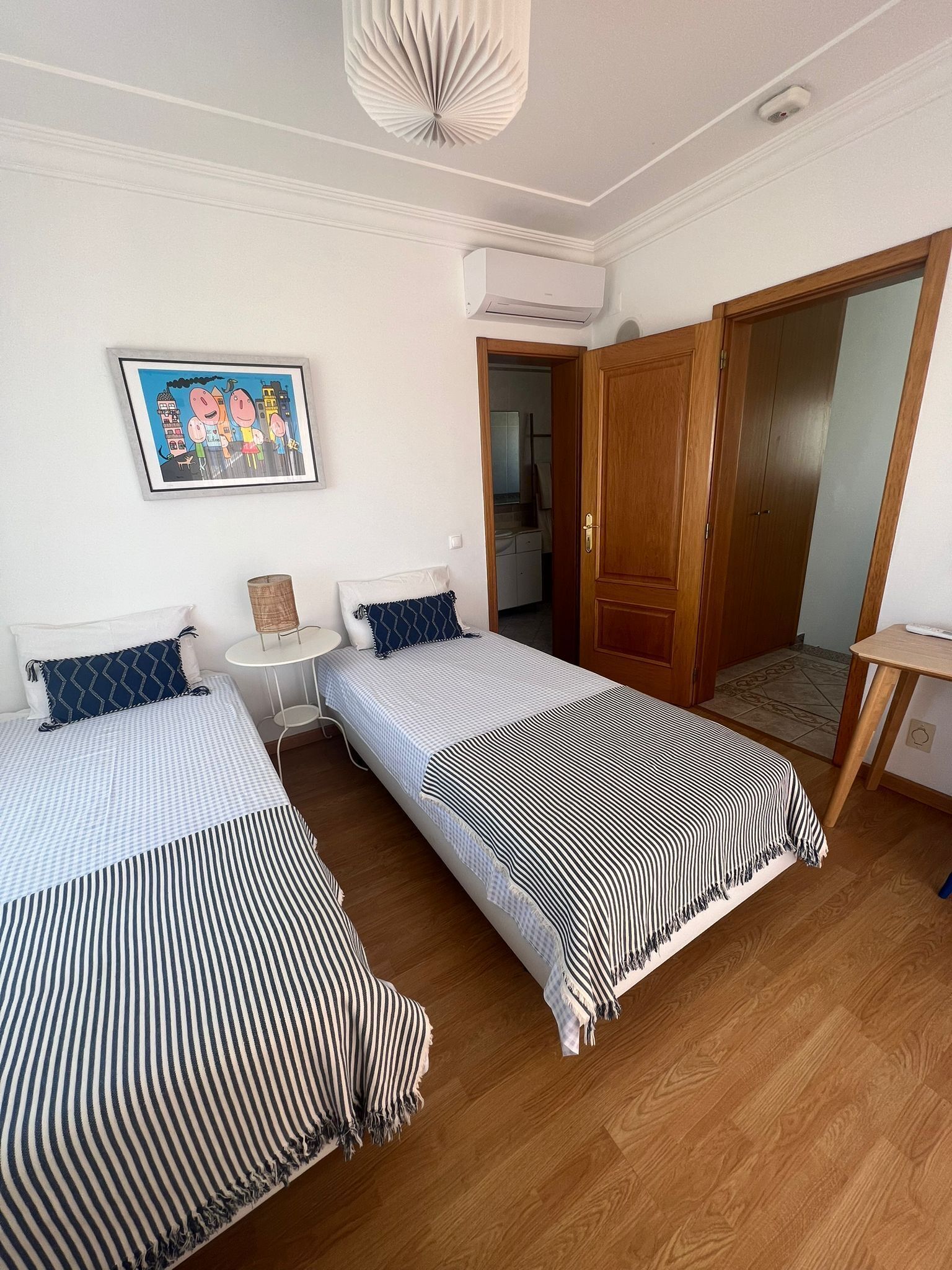 Ein Zimmer mit zwei Betten, einem kleinen Tisch, einer Wand-Klimaanlage und einer Tür zu einem anderen Raum.