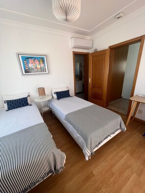 A room with two beds, a small table, a wall-mounted air conditioner, and a door leading to another room. - Villa Med - private pool, garden, barbecue, air conditioning, and ocean view (Olhos de Água)
