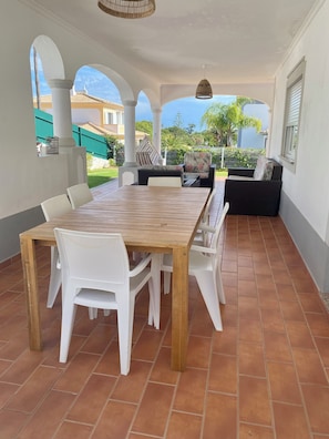 Een overdekt terras met een houten tafel en witte stoelen, een zwarte bank en een hanglamp.