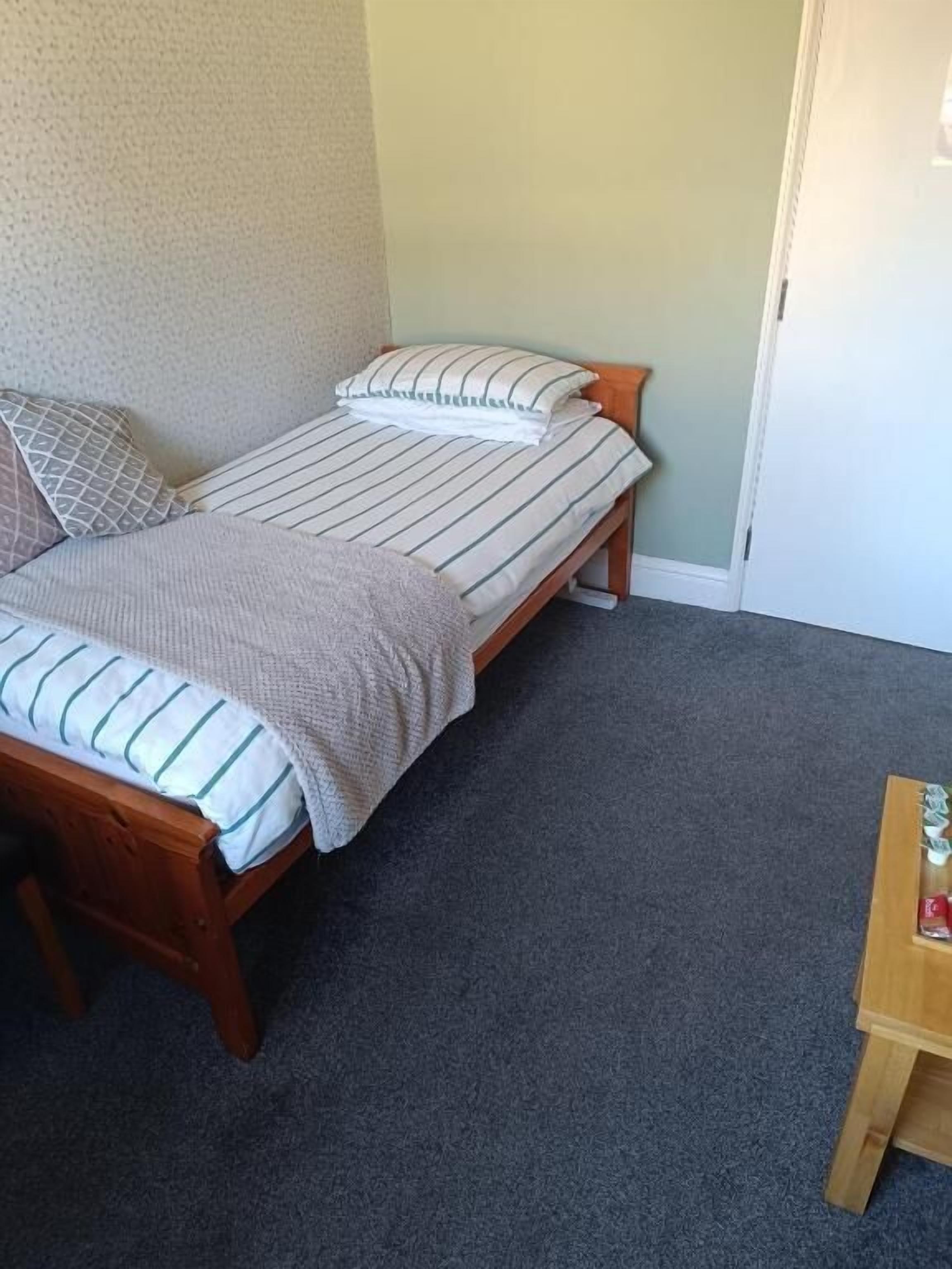 A single bed with a striped bedspread and pillows, a wooden bench, and a small wooden table with a bottle on it.
