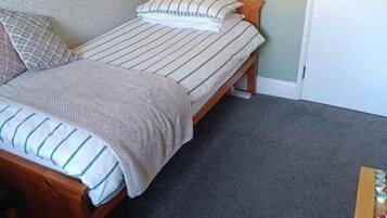 A single bed with a striped bedspread and pillows, a wooden bench, and a small wooden table with a bottle on it.
