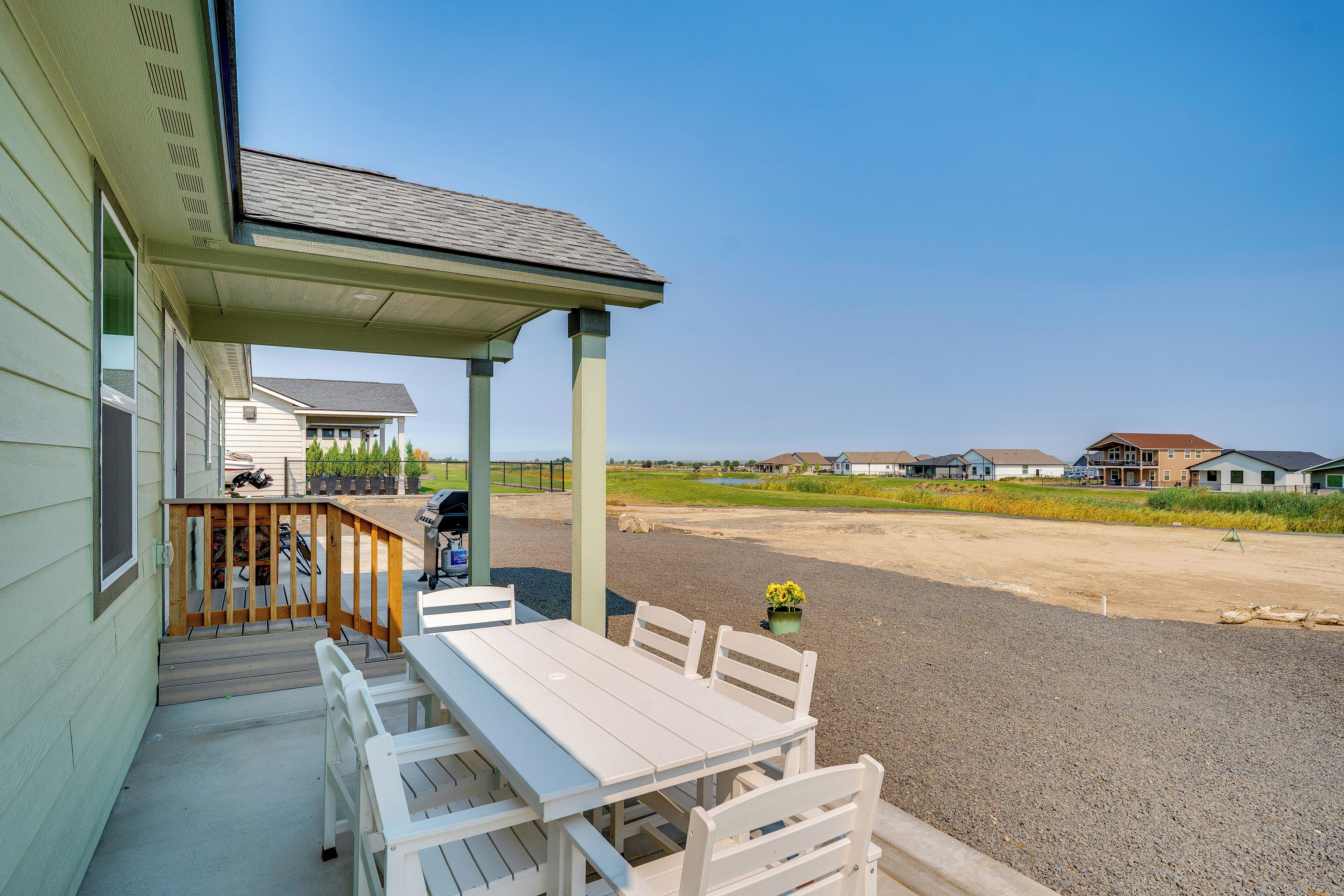 Un espacio al aire libre techado con mesa y sillas, una parrilla y vistas a un barrio residencial con casas y cielo despejado.