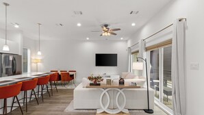A modern kitchen with a dining area, a flat-screen TV mounted on the wall, and a large window with blinds. - Luxurious 3-Bedroom Home In Downtown Houston Area (Houston)