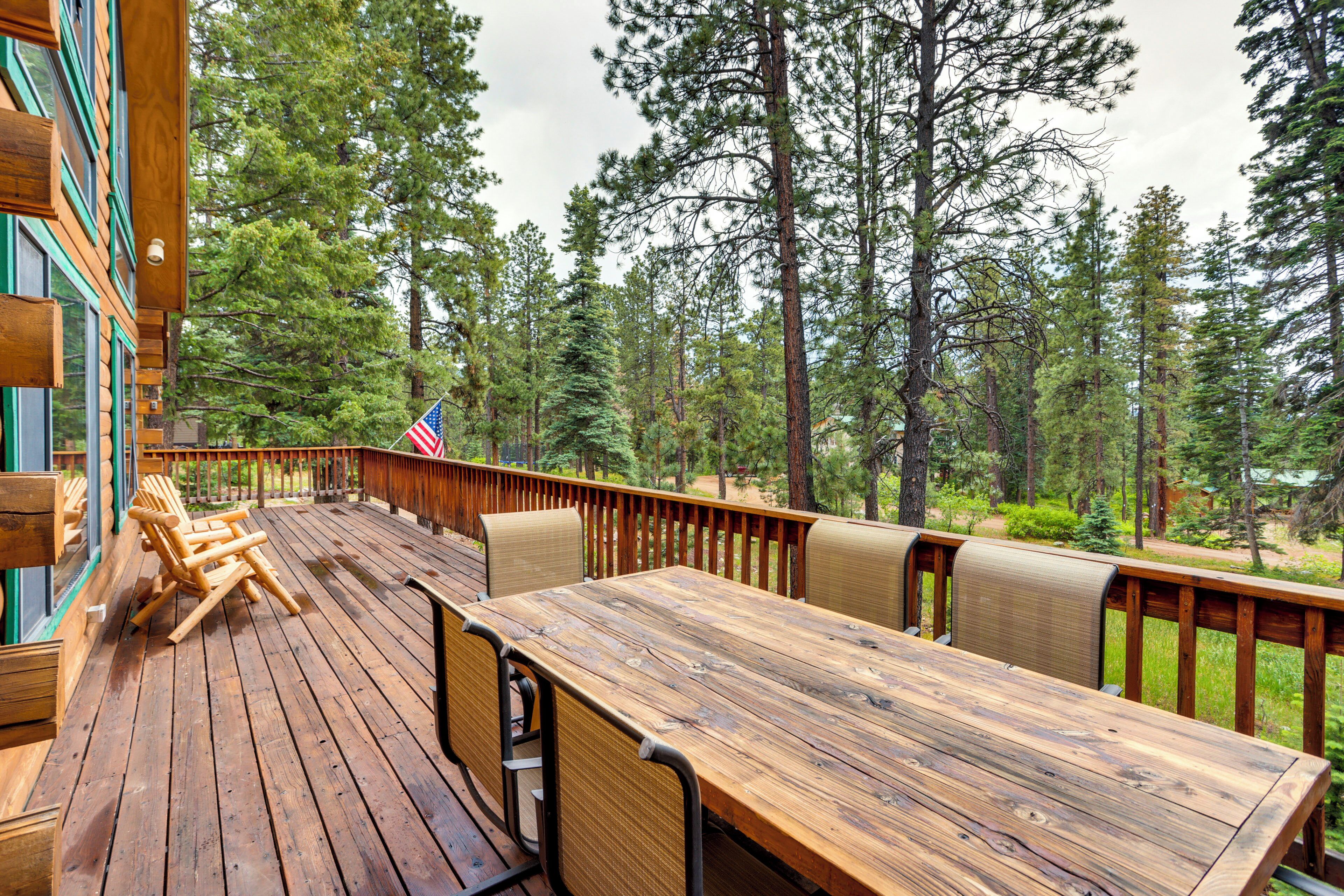 A wooden deck with a table and chairs, surrounded by tall pine trees.