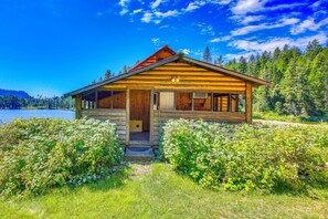 En træhytte med veranda, omgivet af frodig vegetation og en sø.