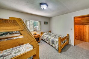 Ein Zimmer mit zwei Holzbettgestellen übereinander, einem Fenster mit Vorhängen, einem kleinen Schreibtisch mit Lampe und einer Holztür.