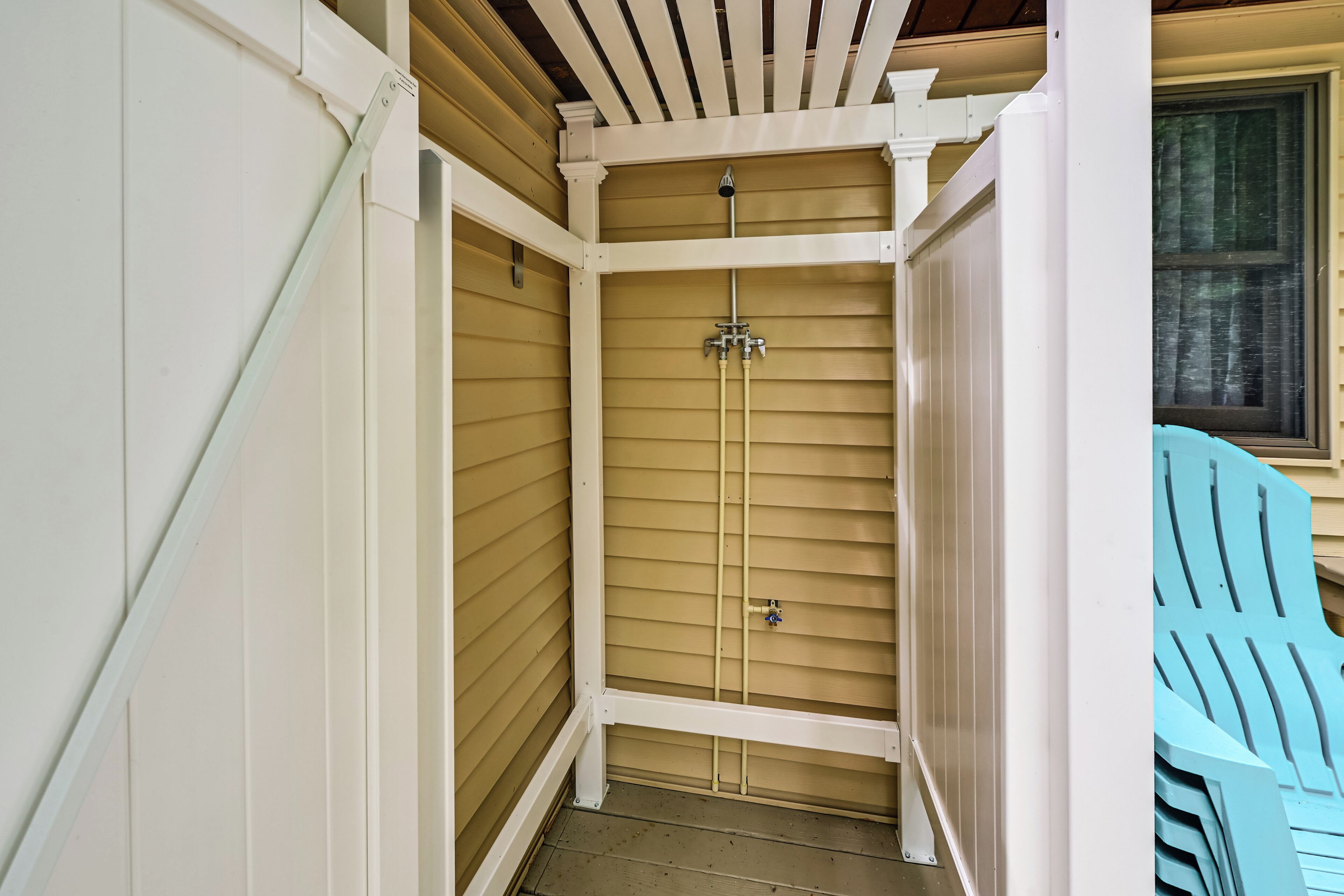 A small, enclosed outdoor shower area with a wooden ceiling and walls, a metal showerhead, and a window to the right.