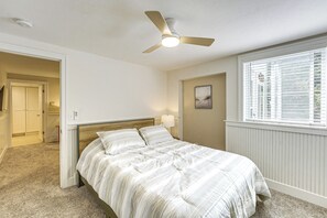 Une chambre à coucher comprenant un lit, un ventilateur de plafond, une fenêtre avec des stores, une porte et un luminaire fixé au mur.