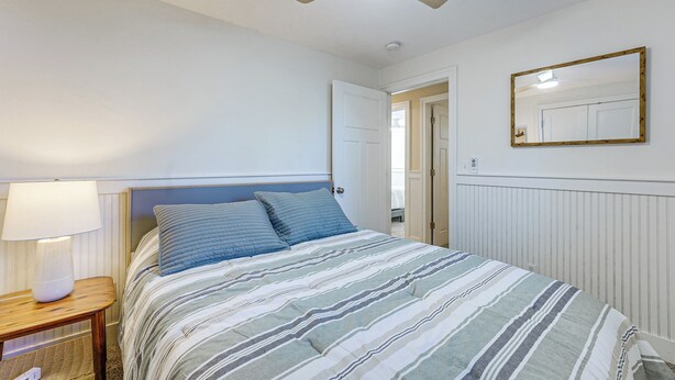 A bedroom with a bed, a bedside table with a lamp, a ceiling fan, and a mirror on the wall.