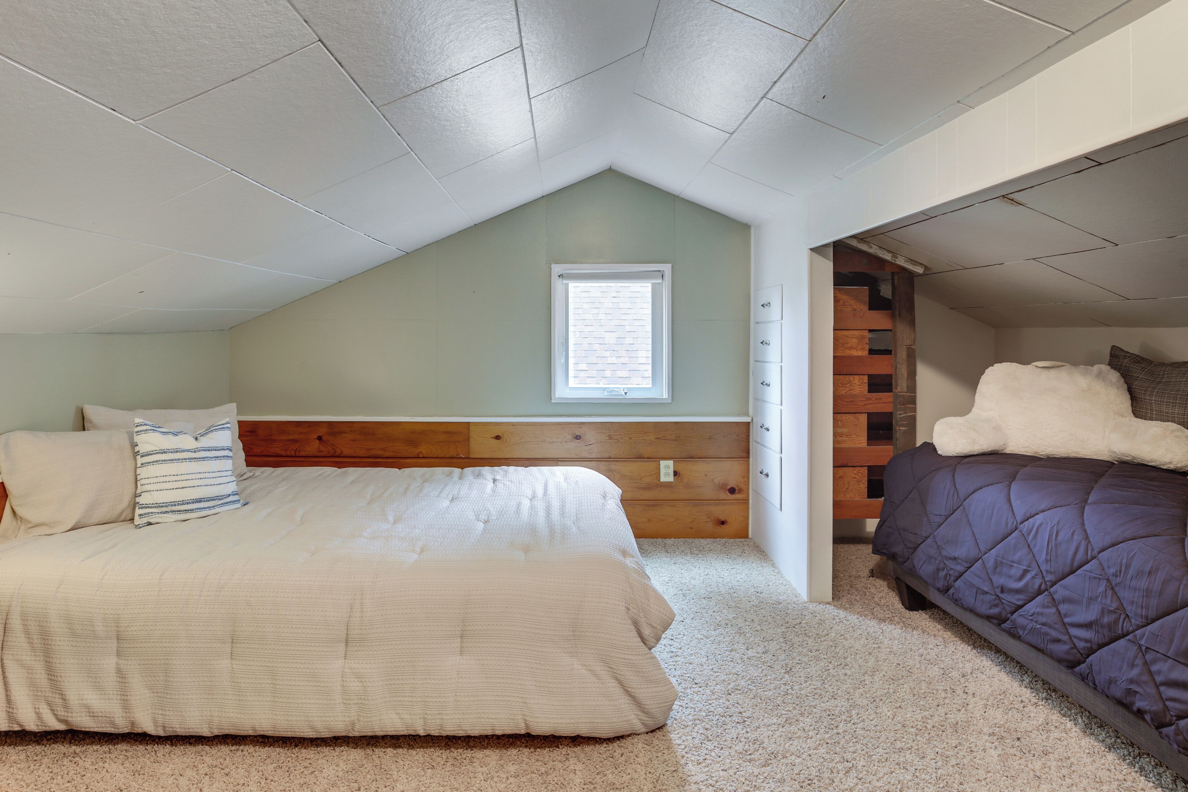 A bedroom with a sloped ceiling, a window, a wooden headboard, a bed with a white comforter, a purple comforter bed, and a small wooden dresser.