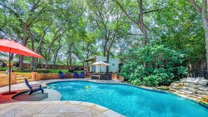 Une piscine avec une terrasse en pierre, entourée d’arbres et d’un petit bâtiment.