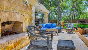 Une terrasse extérieure aménagée avec des meubles en osier, une cheminée en pierre et une vue sur les arbres environnants.