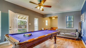 Une table de billard, un ventilateur de plafond, un canapé et une fenêtre avec des stores.