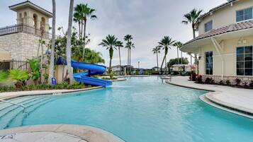 A swimming pool with a blue water slide, surrounded by palm trees and a stone building.
