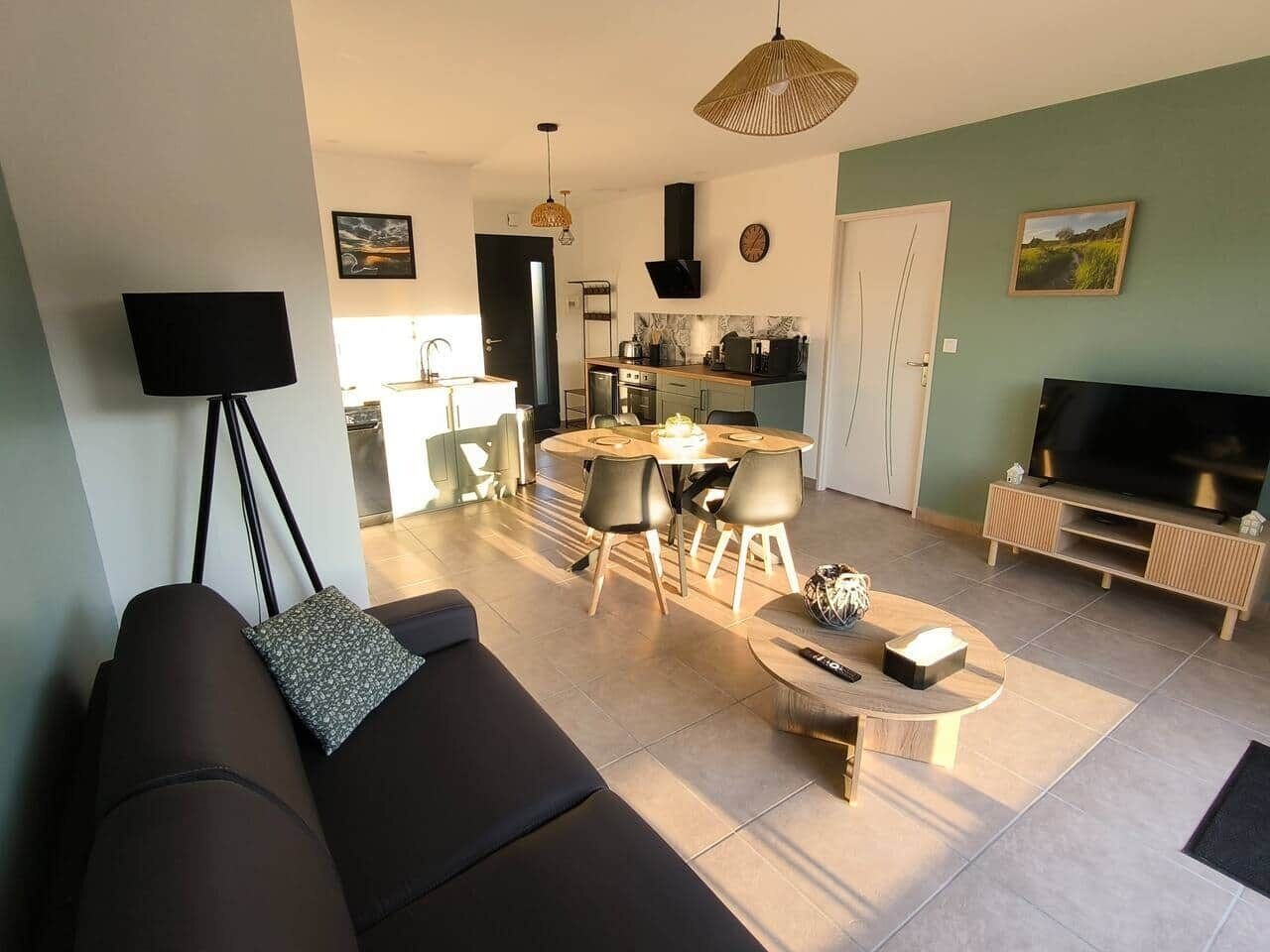 A modern living room with a sofa, a coffee table, and a TV. There is a dining area with chairs and a table. The room has a kitchen in the background.