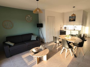 Ein modernes Wohnzimmer mit Sofa, Couchtisch und Essbereich mit Stühlen. Im Hintergrund befindet sich eine Küche mit Spüle und Herd.