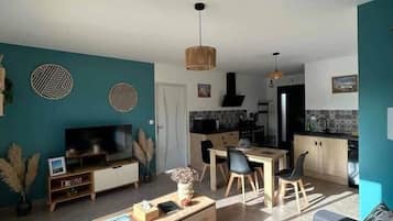A modern living room with a sofa, coffee table, and TV, a dining area with chairs, and a kitchen with a stove and cabinets.