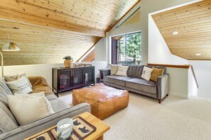 Ein Loft-Wohnzimmer mit hohen Decken, einer grauen Sofagarnitur, einem Lederhocker und einem großen Fenster mit Blick auf Grünflächen.