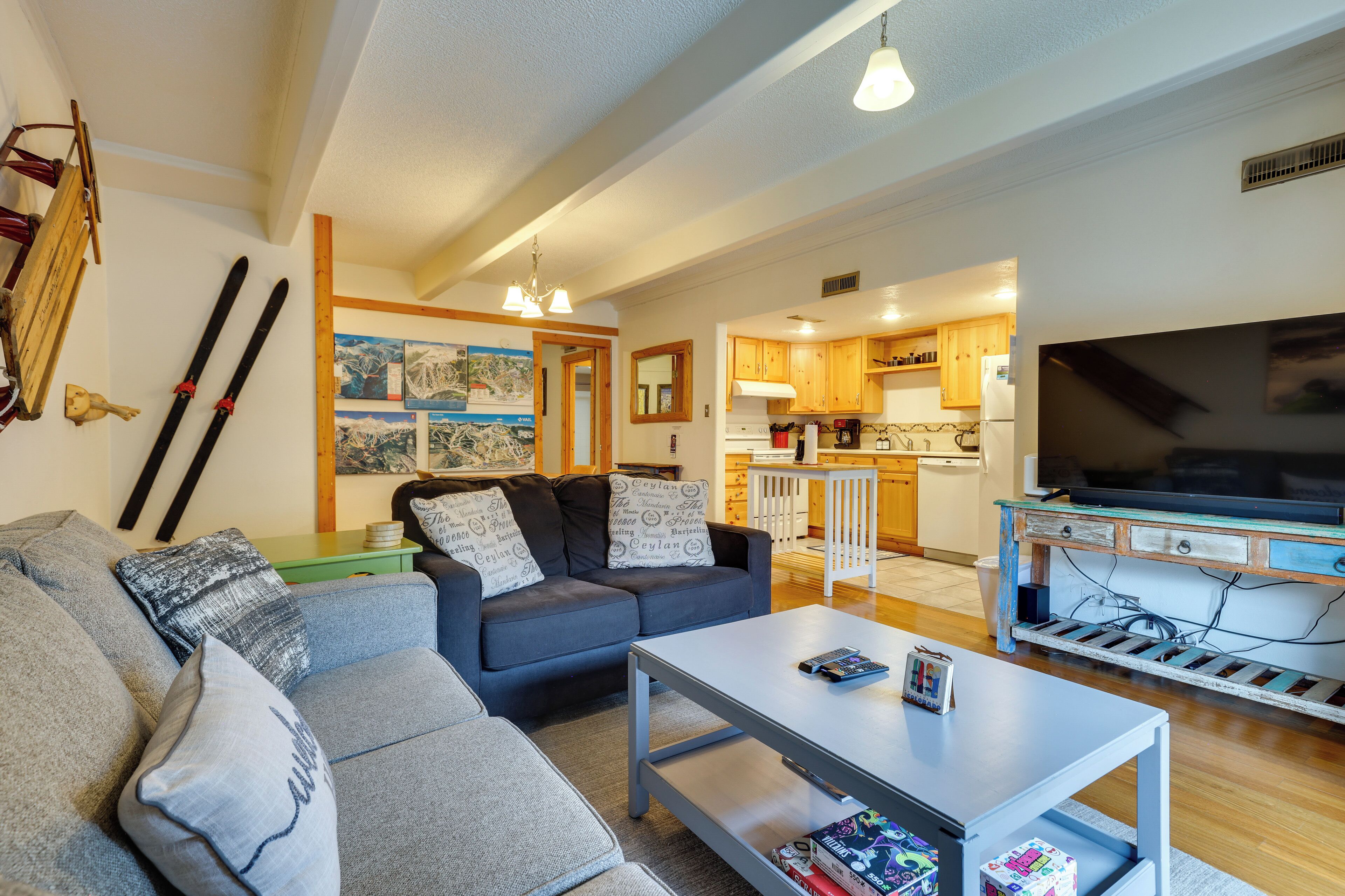 A living room with a sofa, a coffee table, and a television. There is a kitchen area with wooden cabinets and a refrigerator.