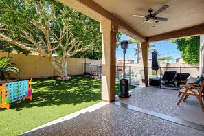 Ein überdachter Patio-Bereich mit Deckenventilator, Sitzmöbeln im Freien, einem Baum und einer Aufstellung für ein Rasenspiel.