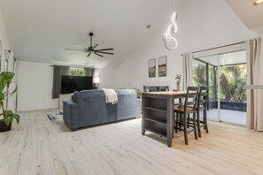 A modern living room with a sofa, TV, ceiling fan, dining table, chairs, and a view of greenery through a glass door. - 4 Mi to Sea Turtle Beach! Family Retreat w/ Yard (Jensen Beach)
