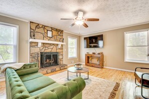 A living room with a stone fireplace, a green sofa, a ceiling fan, and a television mounted on the wall. - Family Getaway in Kennesaw! Spacious Home (Kennesaw)