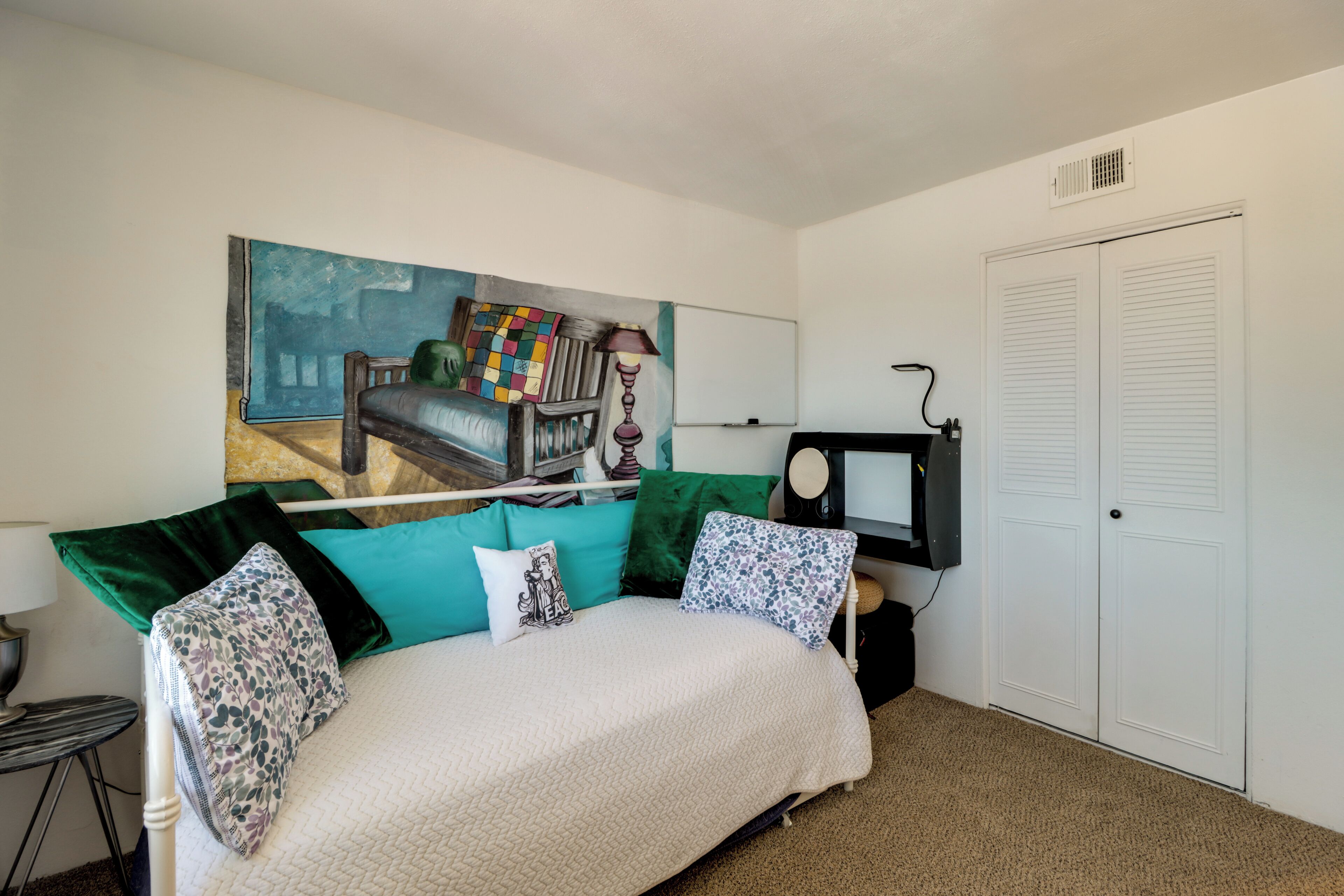 A bedroom with a bed, a painting on the wall, a desk with a computer, and a closet.