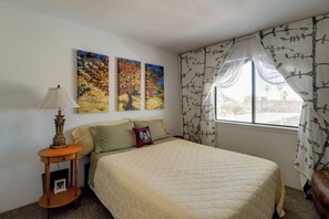 Une chambre à coucher avec un lit, une table de chevet, une lampe, deux tableaux et une fenêtre avec des rideaux.