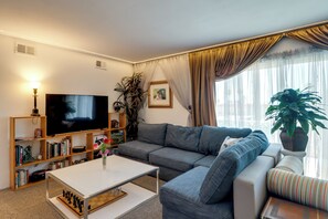Ein Wohnzimmer mit Fernseher, Sofa, Couchtisch, Bücherregal und einem großen Fenster mit Vorhängen.
