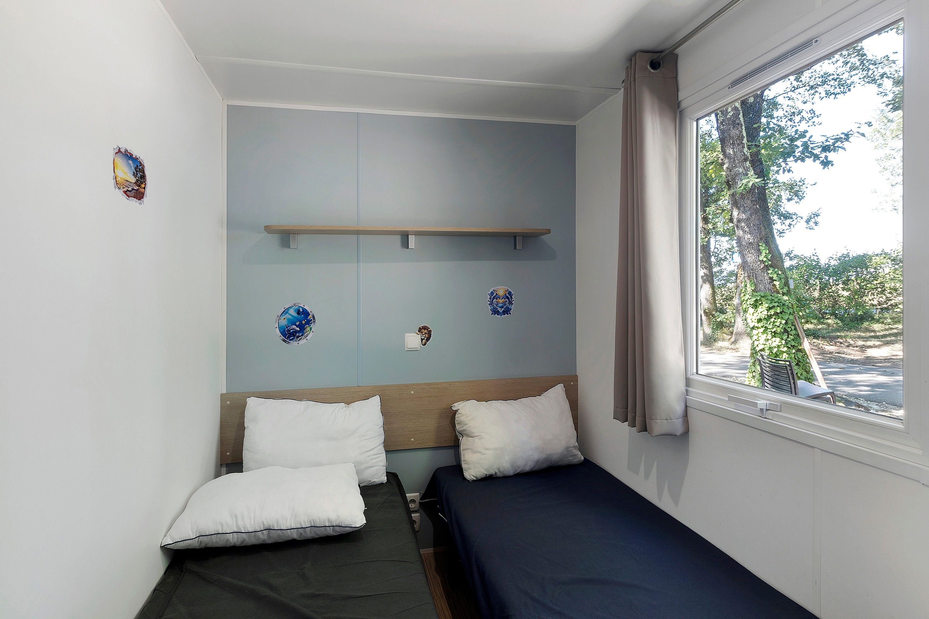 A small, single-bed room with a window, a wooden shelf, and a wall-mounted light switch.