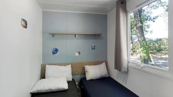 A small, single-bed room with a window, a wooden shelf, and a wall-mounted light switch.