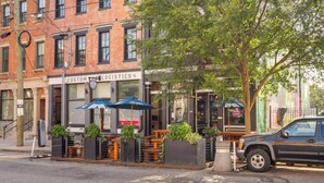 Une vue de rue avec un bâtiment en briques, des tables et des chaises à l’extérieur, protégées par des parasols, et un véhicule garé.