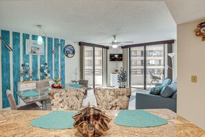 A modern living room with a dining area, large windows, and a ceiling fan. - Dream Daytona Getaway: Beachfront Condo! (Daytona Beach)
