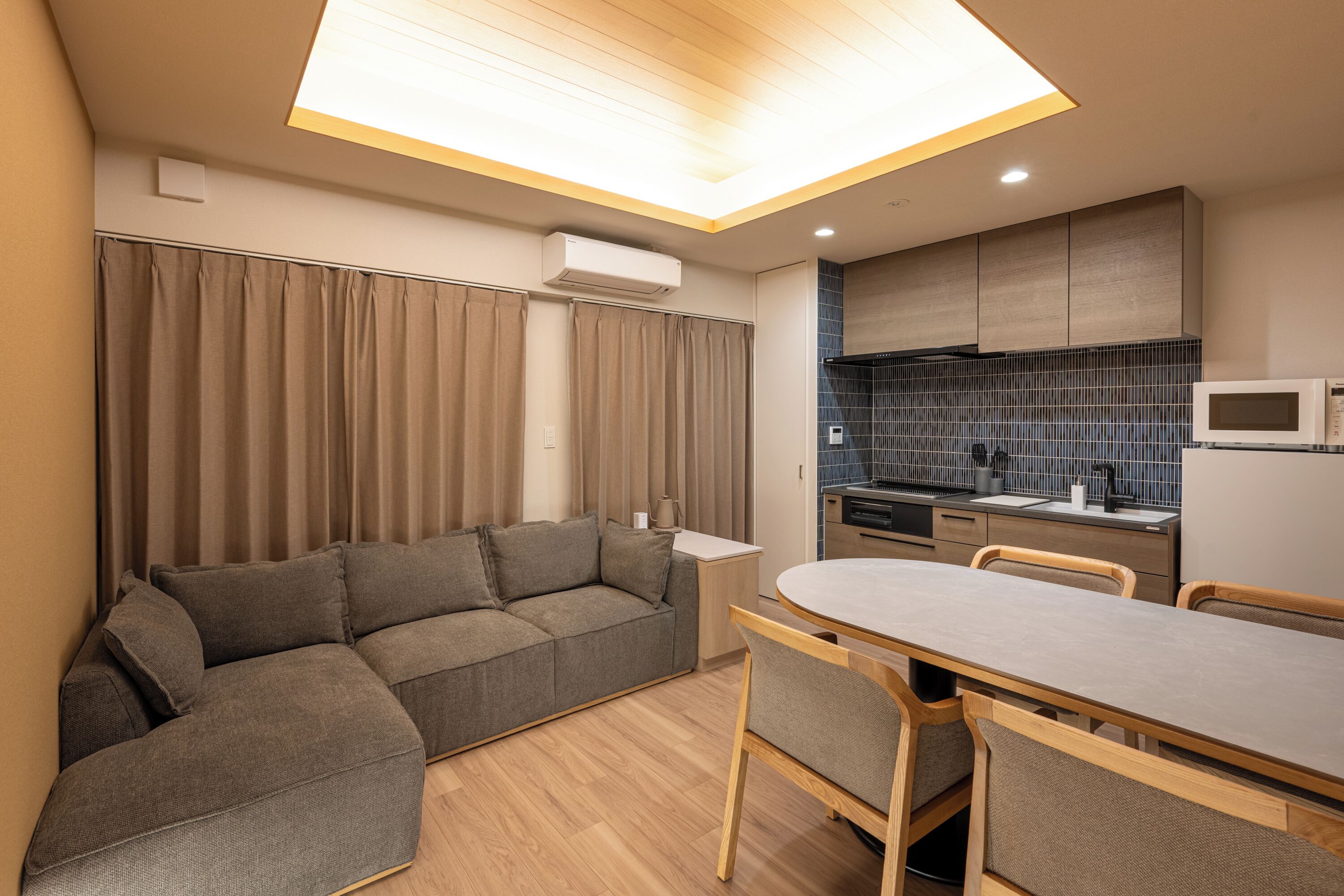 A modern living room with a sectional sofa, a dining table with chairs, and a kitchen area with a microwave.