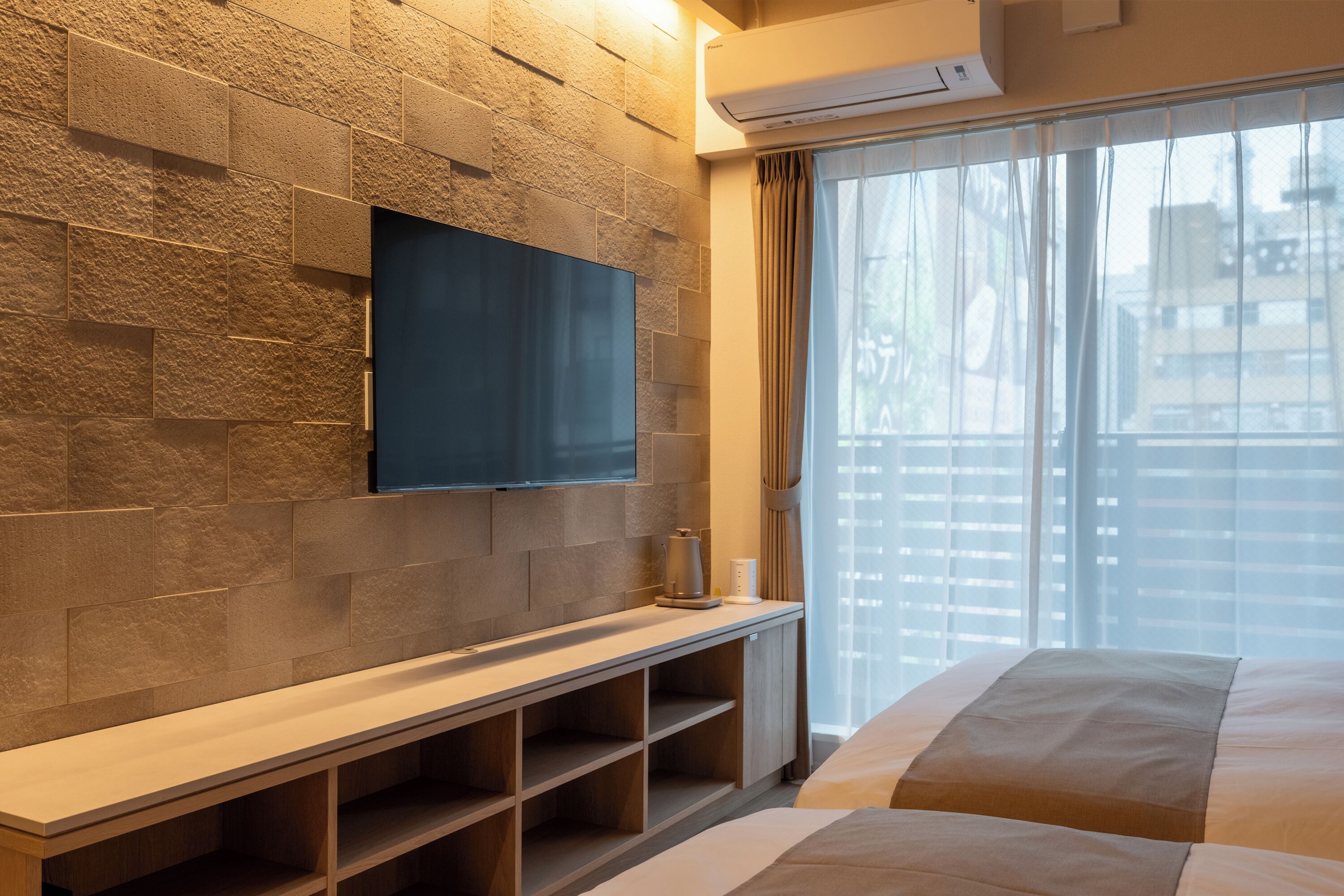 Une chambre d’hôtel moderne avec un mur à la texture de pierre, un téléviseur à écran plat fixé au-dessus d’une console en bois et une grande fenêtre ornée de rideaux légers.