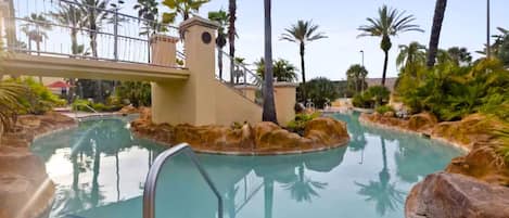 A swimming pool with a stone border, palm trees, and a bridge.