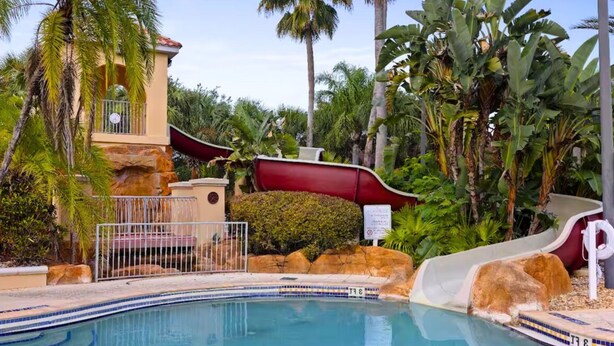Un espace piscine avec un toboggan, des palmiers et un bâtiment doté d’une horloge.