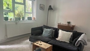 Sala de estar com um sofá preto, mesa de centro de madeira, luminária de chão e janela com plantas.