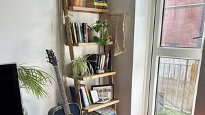 Um quarto com uma estante de livros, um violão, um puff e uma janela com vista para um muro de tijolos.