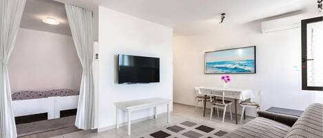 A modern living room with a flat-screen TV, a dining area with a table and chairs, a sofa, and a patterned rug.